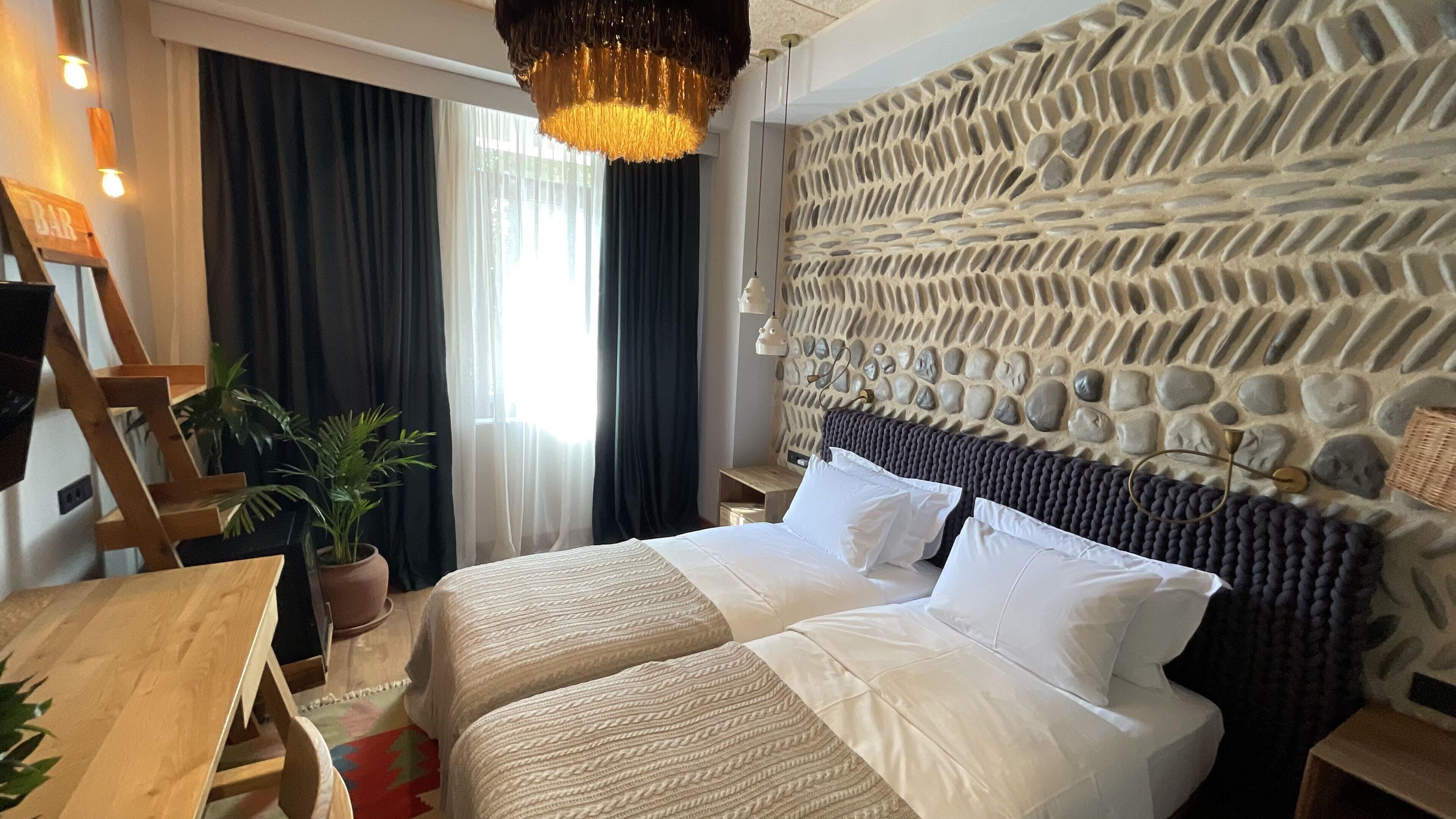 A hotel room with a bed, a desk, a potted plant, and a stone wall.