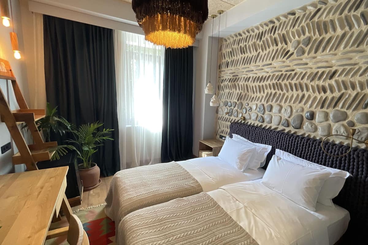A hotel room with a bed, a desk, a potted plant, and a stone wall.