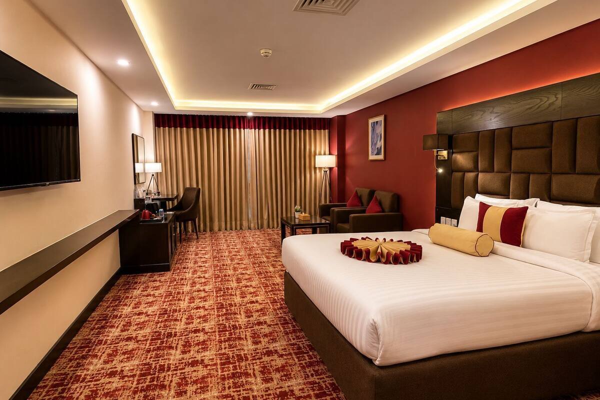 A hotel room with a large bed, a desk, a chair, a TV, and a red carpeted floor.