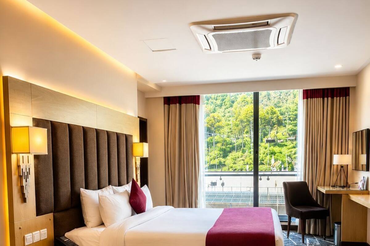 A hotel room with a bed, bedside tables, a desk, a chair, and a large window with a view.