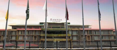 Un hôtel appelé Ramada by Wyndham, doté d’une architecture moderne, de plusieurs mâts de drapeaux et d’une entrée imposante.