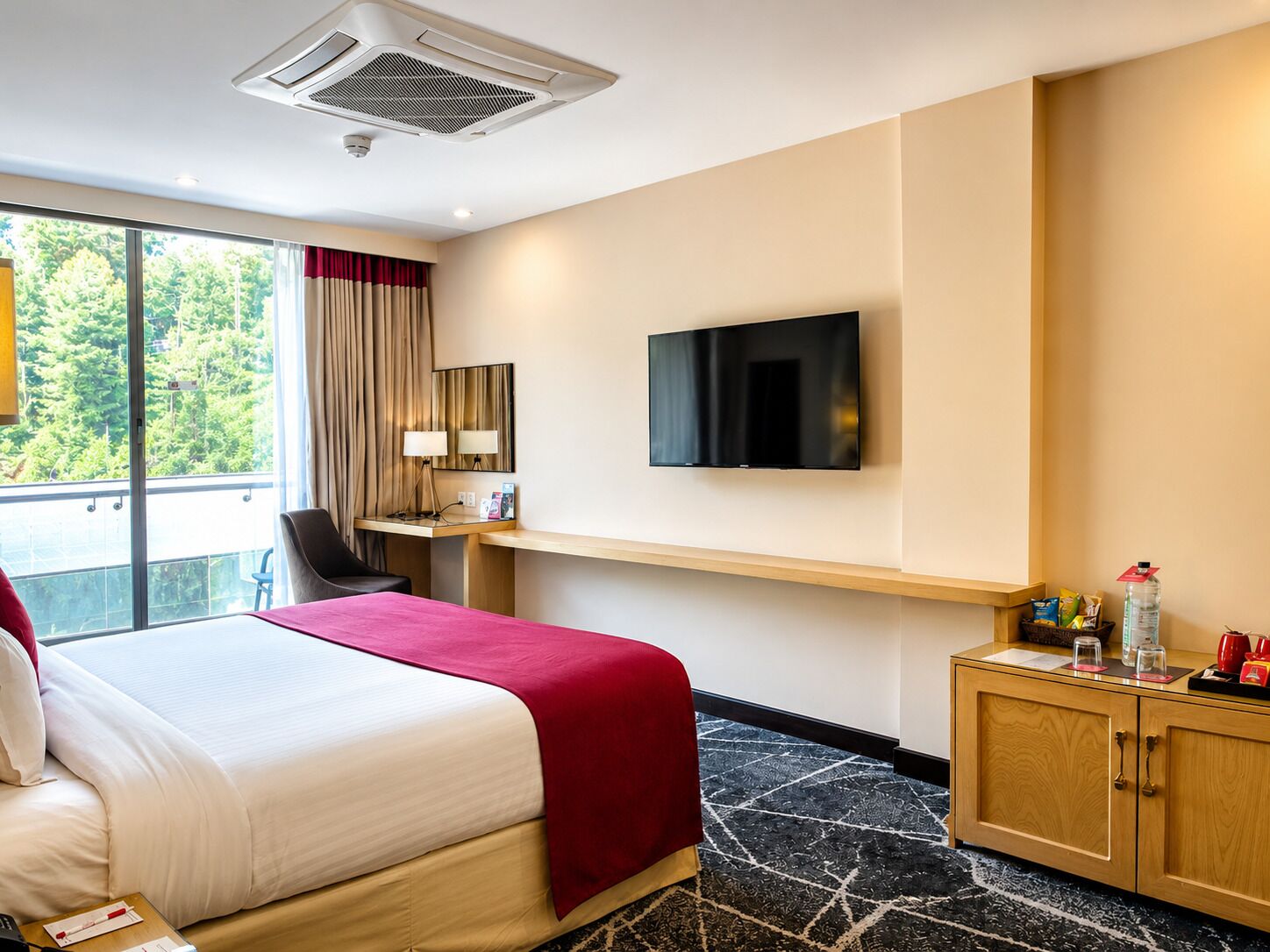A hotel room with a bed, a desk, a TV, a window with curtains, and a view of greenery.