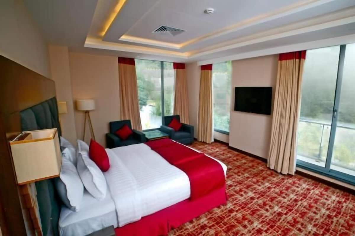 A hotel room with a large bed, two armchairs, a TV, and a balcony with curtains.