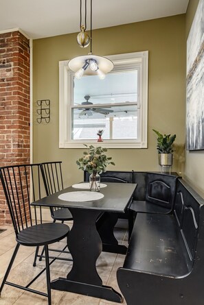Un espace repas avec une table noire et des bancs, un luminaire suspendu, une fenêtre offrant une vue et un mur en briques.