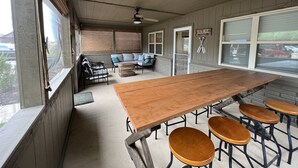 Ein überdachter Patio-Bereich mit einem langen Holztisch und vier Barhocker. Es gibt eine Couch und einen Korbtisch mit Kissen. Ein Schild an der Wand zeigt das Wort „LAKE“.