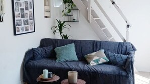Un sofà blau fosc amb nombrosos coixins, dues taules rodones petites amb espelmes, una galeria de fotos emmarcada a la paret, una planta en test i una escala al fons.