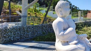 Una escultura de piedra de una figura sentada en un espacio público, con un muro de piedra y follaje al fondo.