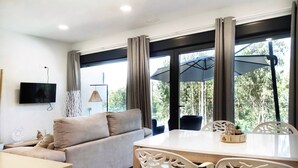 Una sala moderna con un sofá, una mesa de comedor y sillas. Hay un televisor montado en la pared, una lámpara y ventanas grandes con cortinas.