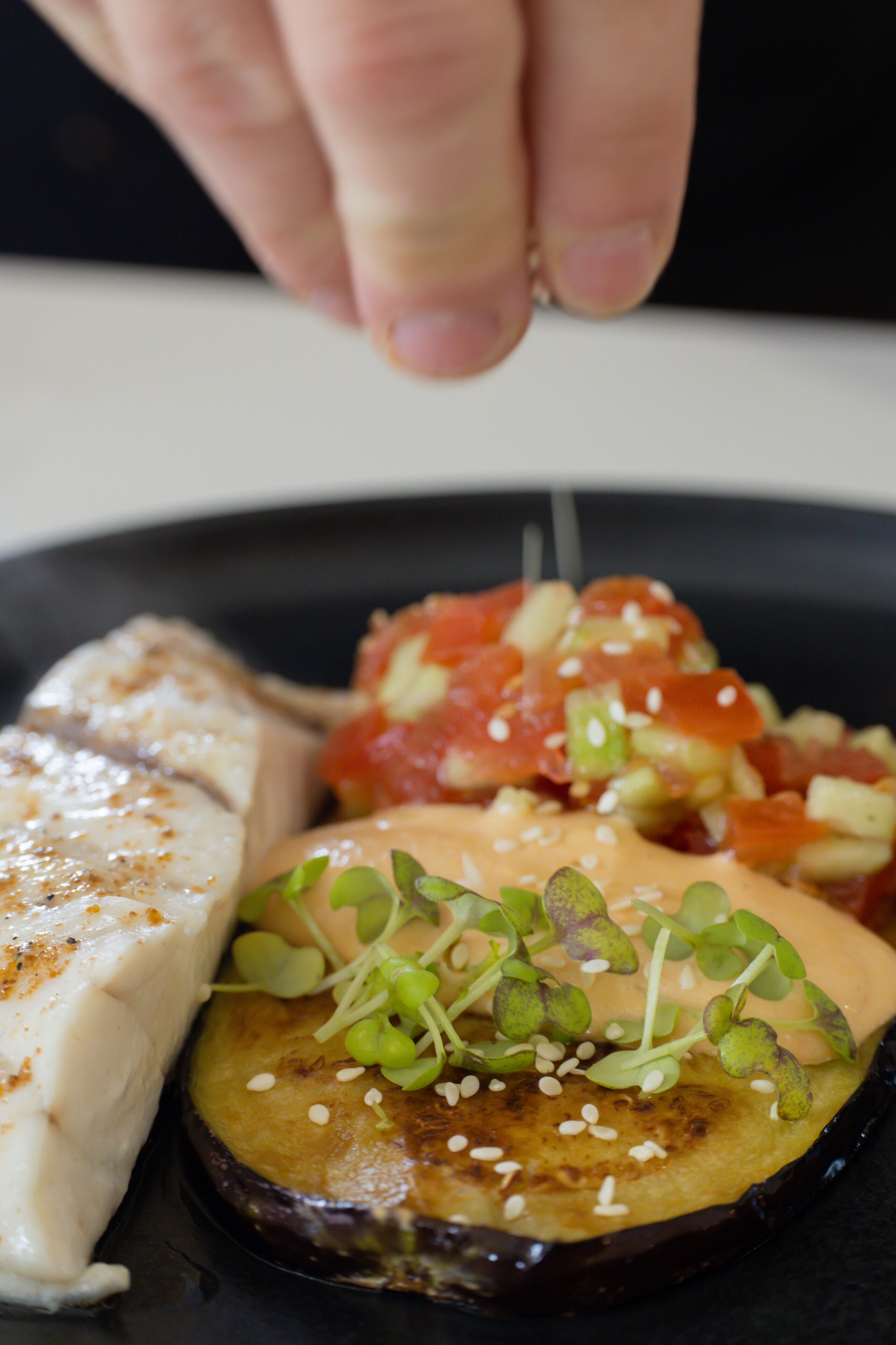 A plate of food with a hand sprinkling sesame seeds.