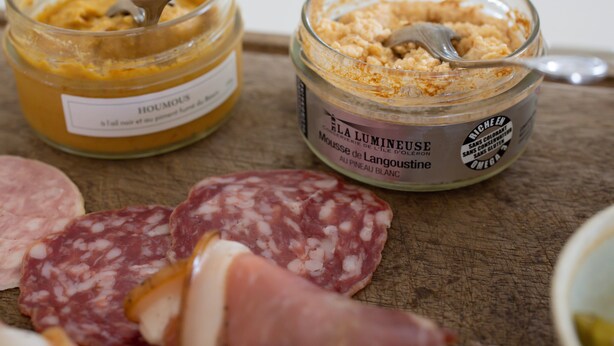 Two jars of food spreads on a wooden surface with slices of salami and a bowl of pickles. - Maison Groleau (Mombrier)