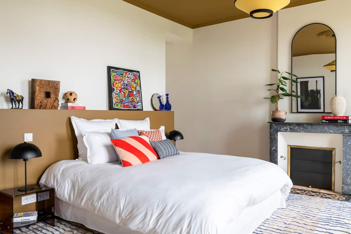 A modern bedroom with a large bed, a nightstand, a wall-mounted shelf with decorative items, a fireplace, and a mirror.