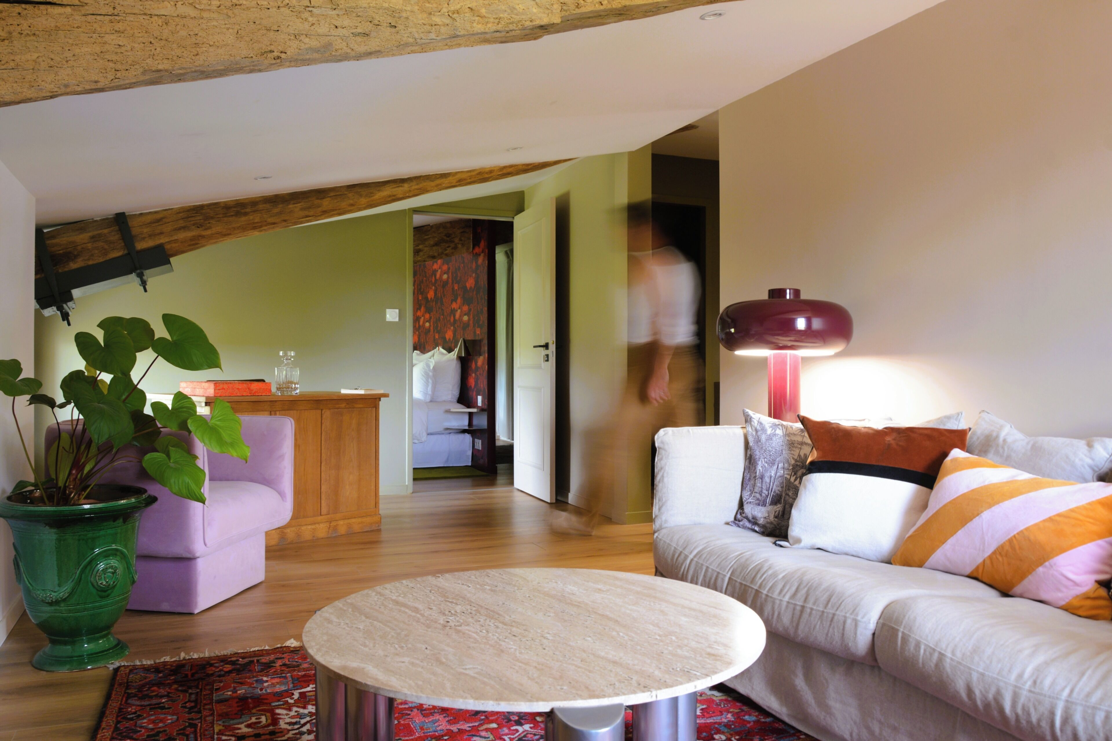A living room with a round table, a sofa with striped pillows, a green plant, and a wooden ceiling beam.