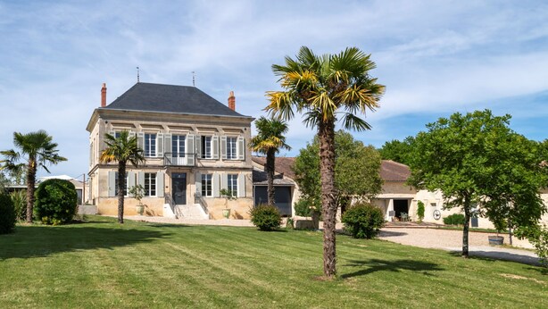 A large, two-story house with a symmetrical facade, multiple windows, and a well-maintained lawn with palm trees. - Maison Groleau (Mombrier)