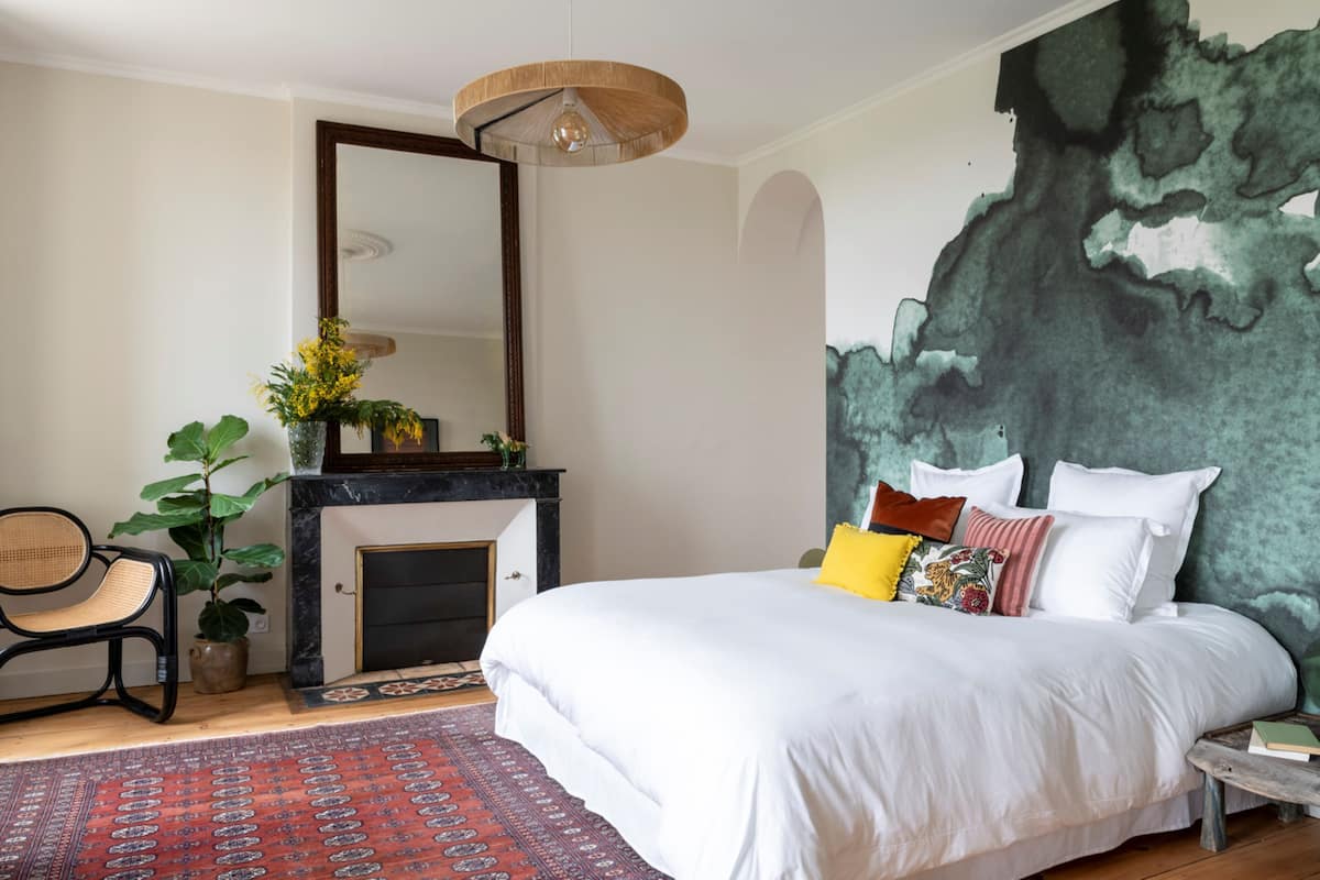 A bedroom with a large bed, a fireplace, a mirror, and a wall mural.
