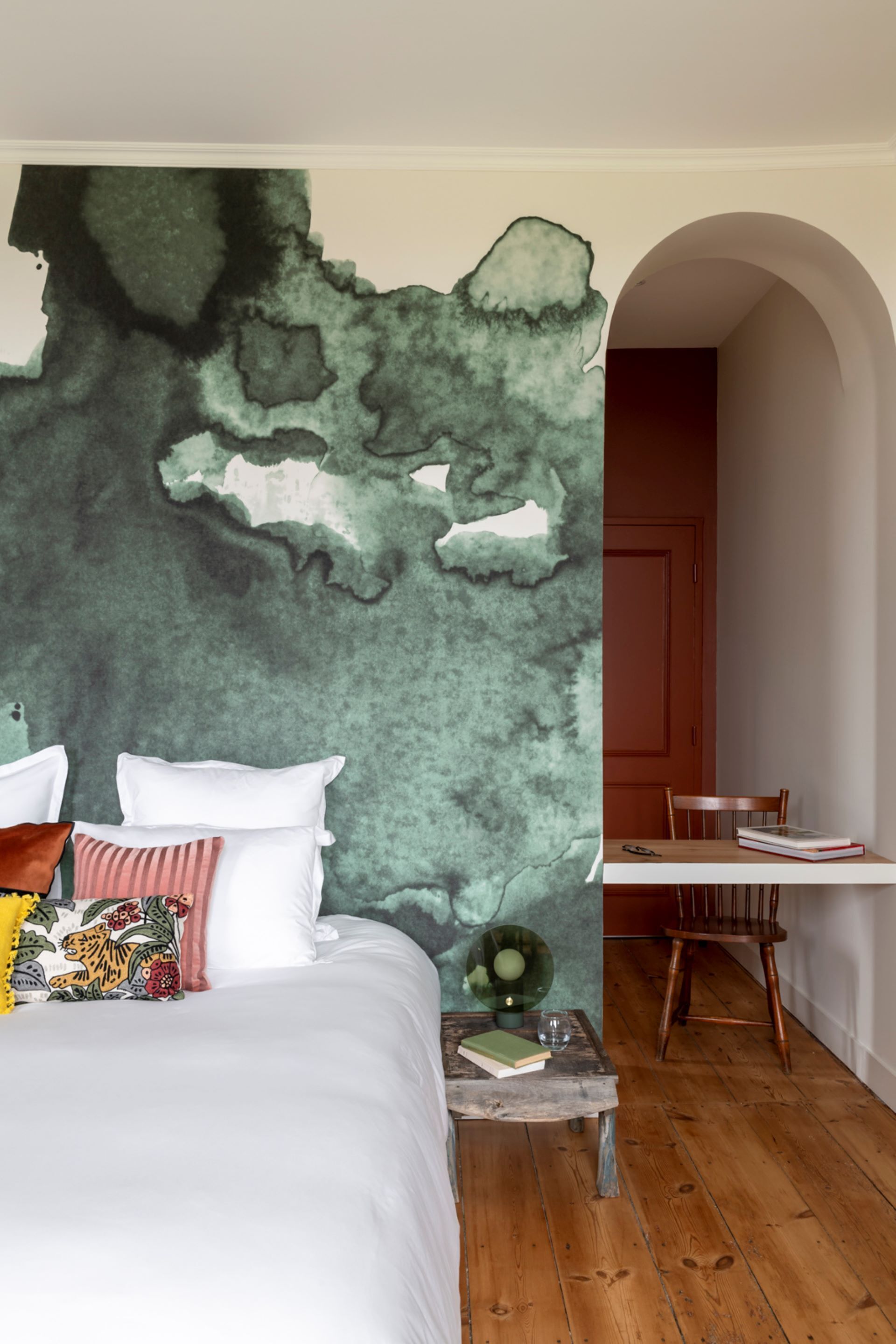 Une chambre avec une grande fresque abstraite sur le mur, un lit avec une literie blanche, une table basse en bois avec des livres et un verre, et un coin repas avec une table et des chaises.