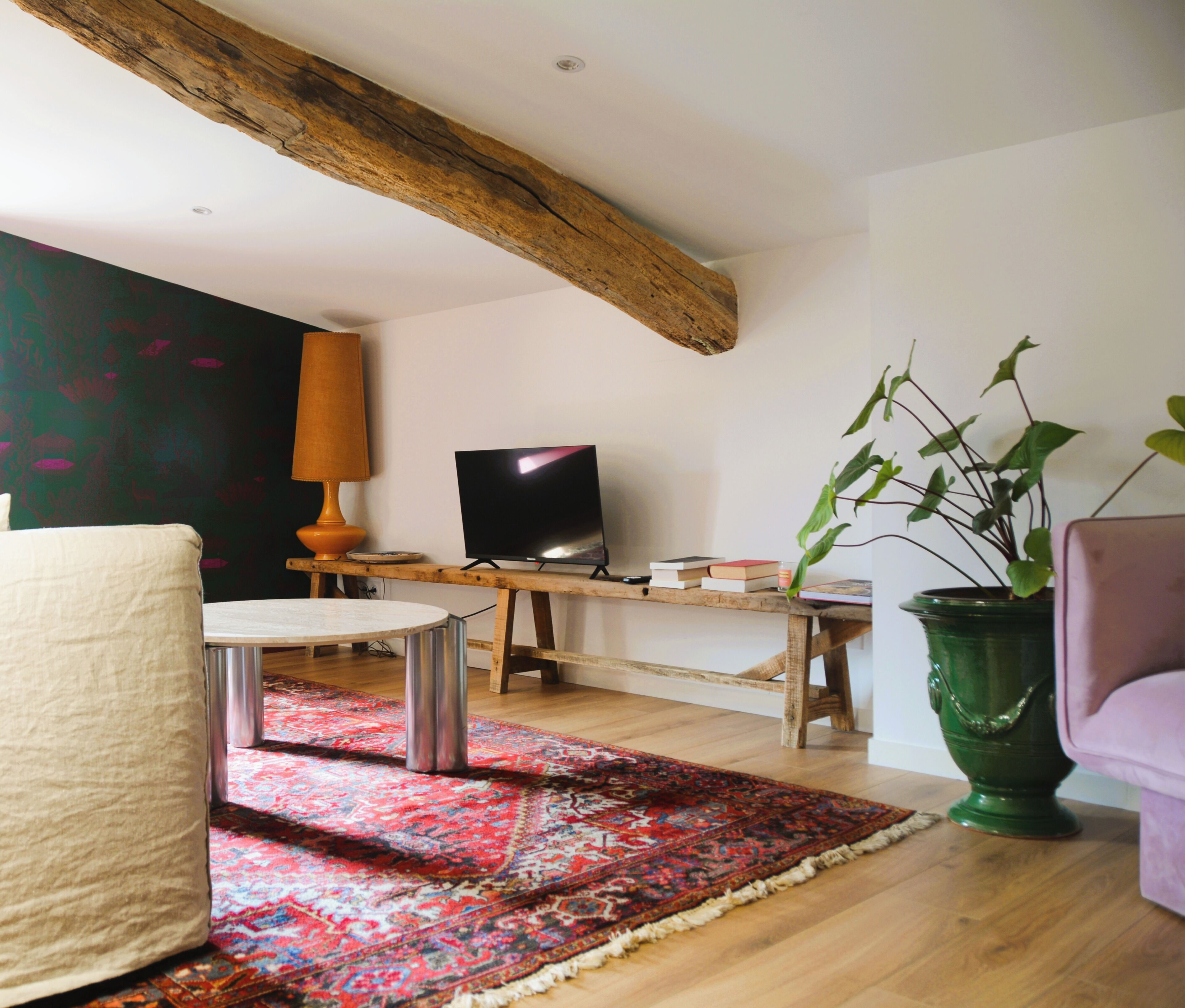 Un salón con techo de madera, televisor, planta verde y alfombra estampada.