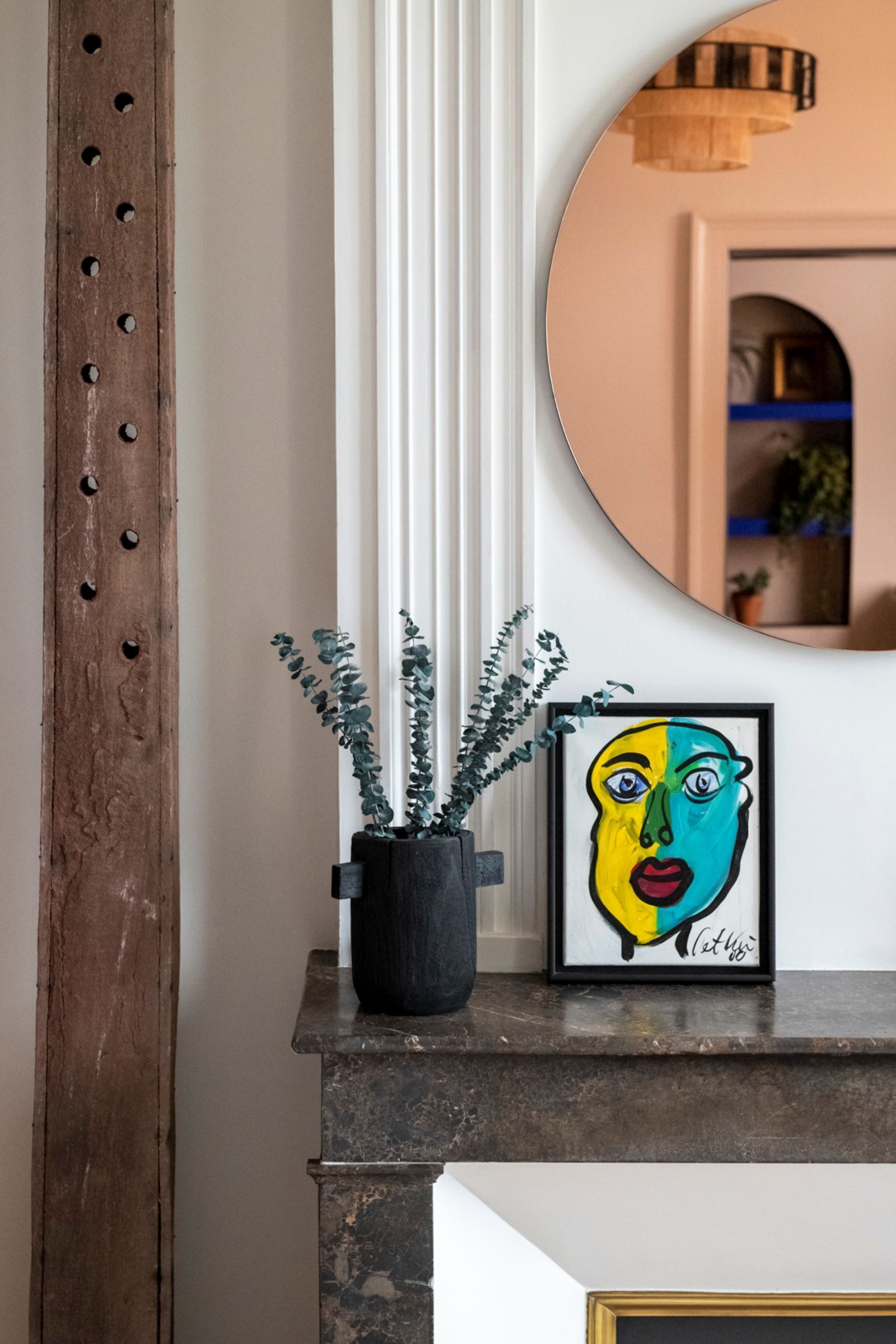 A framed artwork with a blue and yellow face, a potted plant, and a mirror on a mantelpiece.