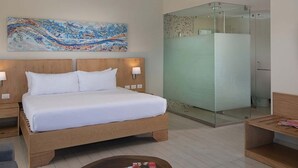 A hotel room with a wooden bed, a glass shower enclosure, and a large abstract painting on the wall. - Luxurious 1 Bedroom Tropical Junior Suite (Dominican Republic)