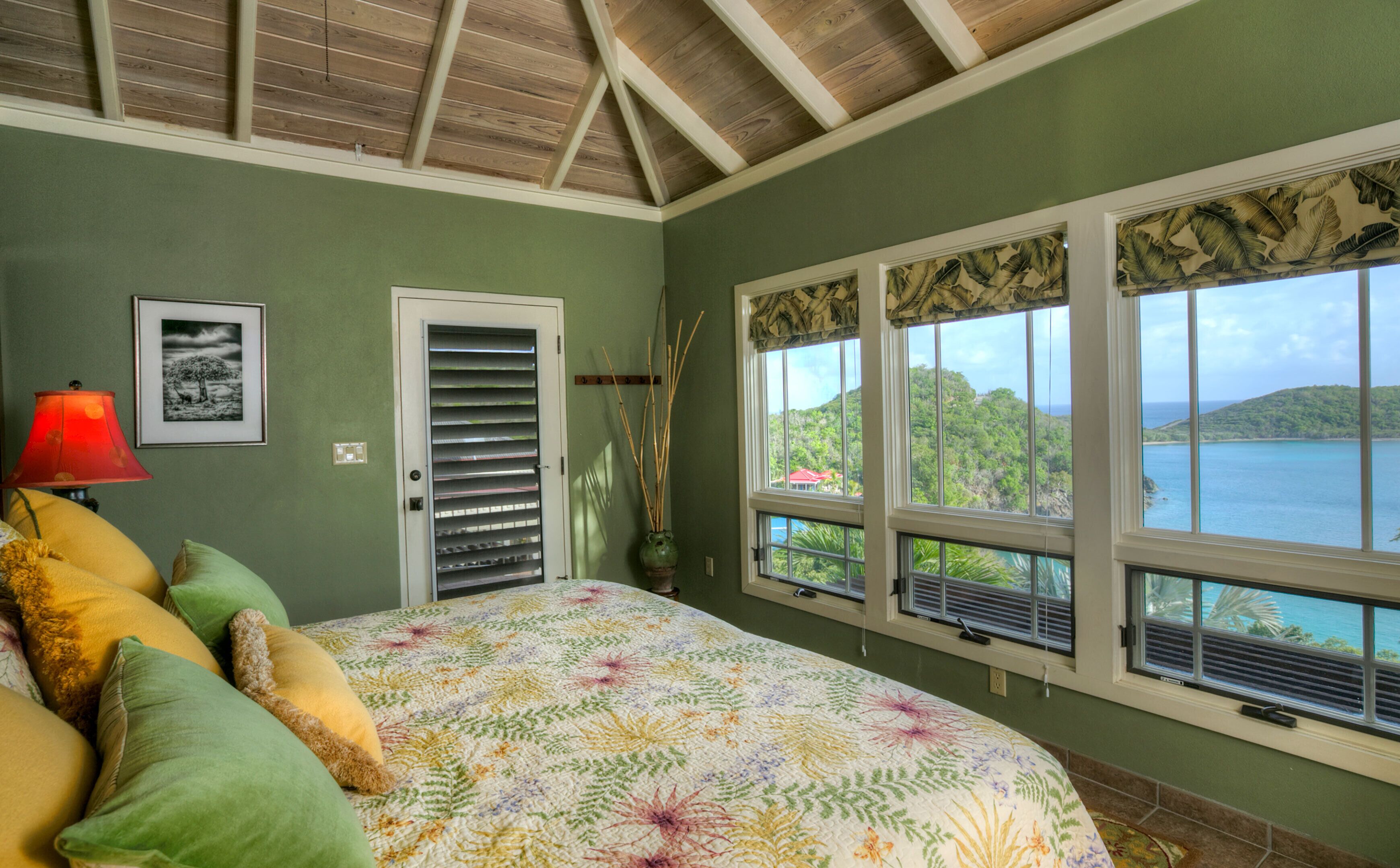 A bedroom with a bed, a window with a view of the sea, a door, and a lamp.