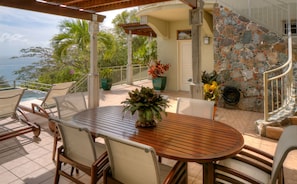 A patio with a round wooden table and chairs, surrounded by potted plants and overlooking a scenic view.