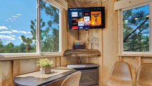 A cozy cabin interior with a wooden interior, a TV mounted on the wall displaying various streaming services, a small table with chairs, and a window offering a view of trees. - Mountainside Tree House Retreat (Larkspur)