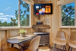 A cozy cabin interior with a wooden interior, a TV mounted on the wall displaying various streaming services, a small table with chairs, and a window offering a view of trees. - Mountainside Tree House Retreat (Larkspur)