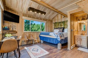 A bedroom with a bed, a desk, chairs, a TV, and a window with a view of trees. - Mountainside Tree House Retreat (Larkspur)