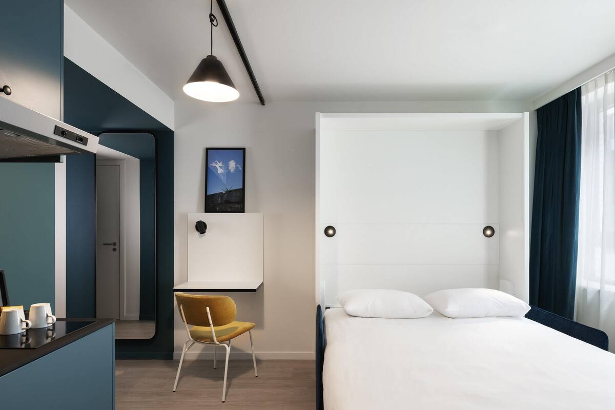 Une chambre d’hôtel moderne, équipée d’un lit, d’un bureau, d’une chaise, d’un tableau au mur et d’un luminaire suspendu.