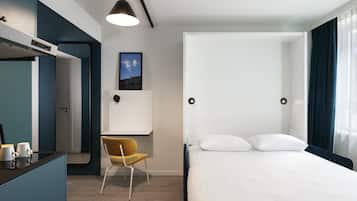 A modern hotel room with a bed, a desk, a chair, a picture on the wall, and a hanging light fixture.