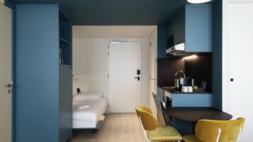 A compact room with a bed, two chairs, a kitchenette, and a door leading to another room.
