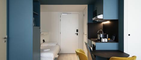 A compact room with a bed, two chairs, a kitchenette, and a door leading to another room.