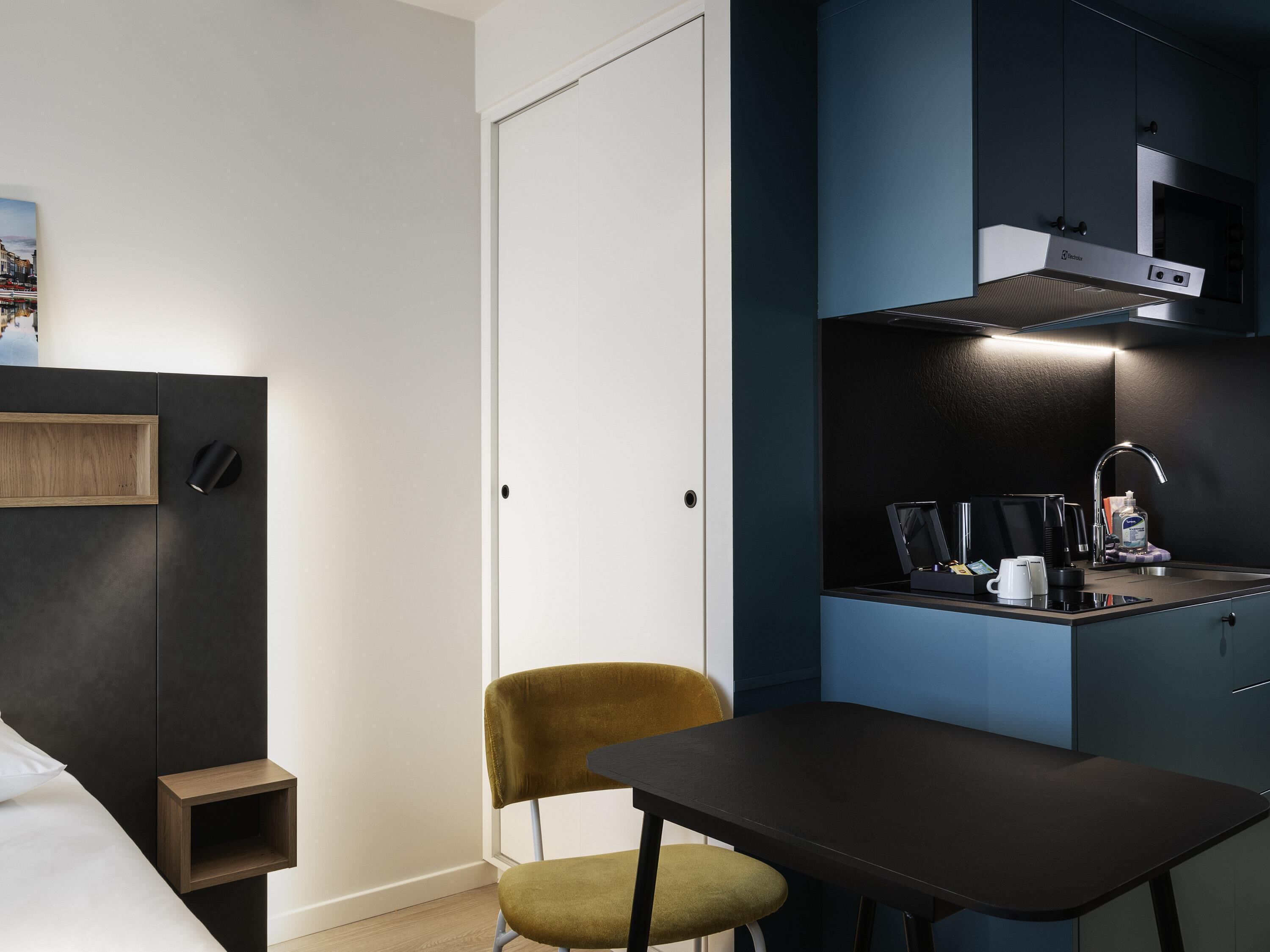 Habitación pequeña con cama, mesita de noche, una silla y una kitchenette equipada con estufa y fregadero.