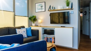 Un salon moderne avec un canapé gris, un fauteuil bleu, un téléviseur à écran plat fixé à un mur blanc, une bibliothèque remplie de livres et une plante en pot.