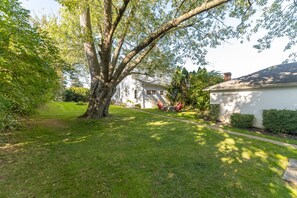 Una propiedad residencial con un gran árbol, césped cuidado y una casa con porche delantero y ventanas visibles.