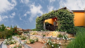 A house with a stone retaining wall, white flowering plants, and a covered patio area. - Large outdoor with privacy, Heated pool 60m2 , Gamesroom ,Wellness, Garden (Karojba)