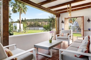 Un porche cubierto con muebles blancos, una mesa y vistas a un paisaje verde.