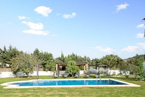Una piscina con sillas de descanso, una pérgola y un jardín con árboles y arbustos.