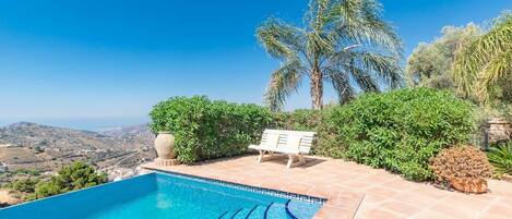 Una piscina con un banco, rodeada de follaje y palmeras, con vistas a un paisaje montañoso.