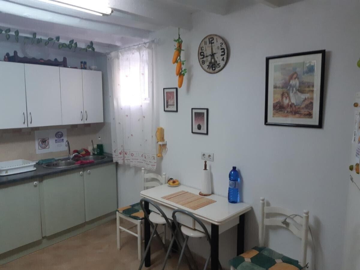 A small kitchen with white cabinets, a dining table with chairs, a clock, and a framed picture on the wall.