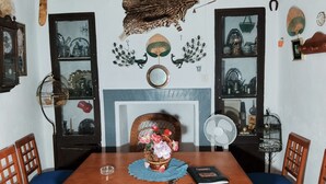 Un coin repas avec une table en bois, des chaises à l’assise rembourrée de couleur bleue, un vase de fleurs, un ventilateur et des éléments de décoration murale, notamment un tableau encadré et un élément décoratif suspendu au mur.