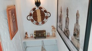 Un couloir étroit, éclairé par une lanterne, où l’on aperçoit une statue de Bouddha, un tableau encadré, une petite commode et un sac suspendu.