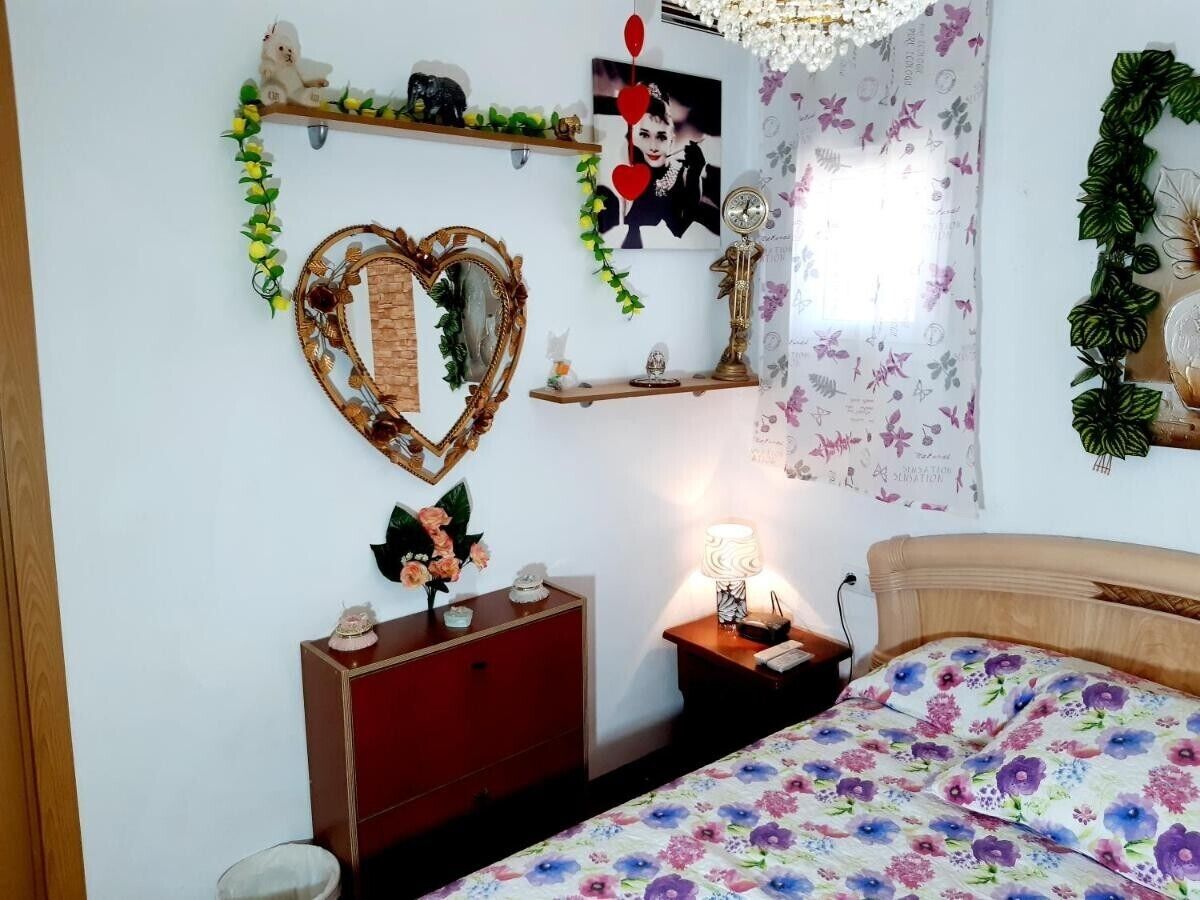 Ein Schlafzimmer mit Bett, Schminktisch, Nachttisch, Wanddekorationen und einem Fenster.
