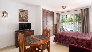 A room with a bed, a desk with a television, a dining table, and a painting on the wall. - Studio Apartment '19A Claire' with Pool, Private Garden & Wi-Fi (El Paraiso)