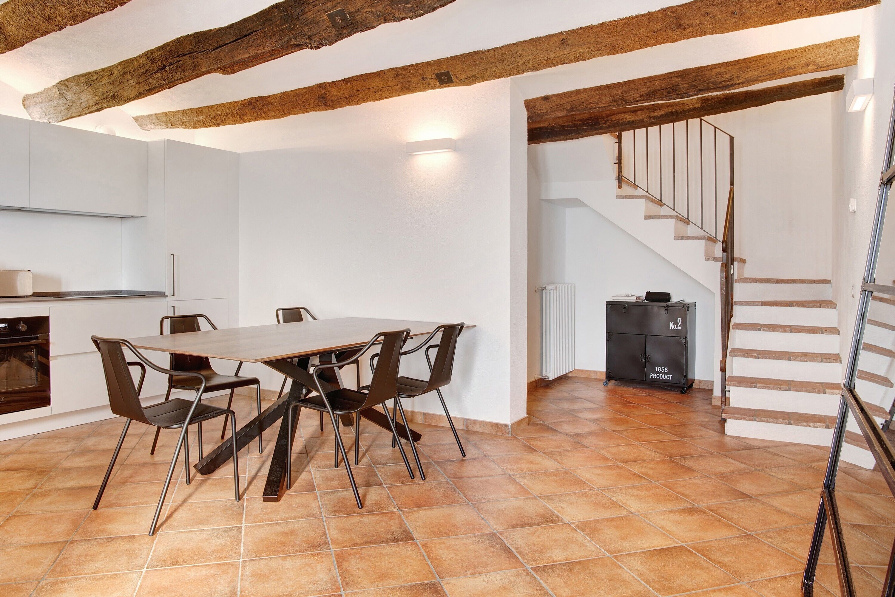 A dining area with a wooden table and chairs, a kitchen with white cabinets, and a staircase leading to an upper level.