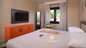 A hotel room with a bed, a TV mounted on the wall, an orange cabinet, and a window with curtains. - Luxurious 1 Bedroom Royal Suite (Dominican Republic)