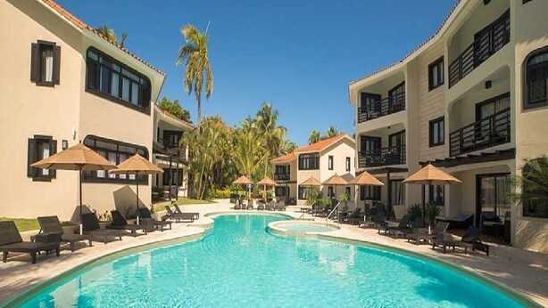A pool area with lounge chairs and umbrellas, surrounded by multi-story buildings with balconies. - Luxurious 1 Bedroom Royal Suite (Dominican Republic)