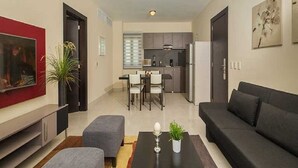 A modern living room with a sofa, a coffee table, and a TV. There is a kitchen in the background with cabinets and a refrigerator. - Luxurious 1 Bedroom Royal Suite (Dominican Republic)