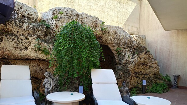 Espace extérieur aménagé avec des coussins blancs et des tables rondes, le tout adossé à un mur à la texture intéressante, agrémenté de verdure.