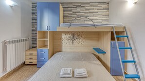 Un lit superposé avec une échelle, un bureau et une commode dans une pièce équipée d’un radiateur et d’une étagère fixée au mur.