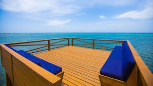 A wooden deck with blue cushioned seating facing a turquoise sea under a clear sky. - Lawson Rock - Yellowfish 211 2 Bedroom Condo by RedAwning (Roatan)